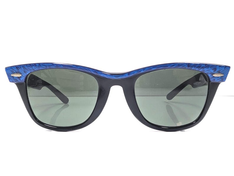 Op de afbeelding: Vintage zonnebril met een zwart montuur en groene glazen. De bovenkant van het montuur heeft een blauw gemarmerd ontwerp. De zonnebril heeft een klassieke cat-eye vorm, op een witte achtergrond.