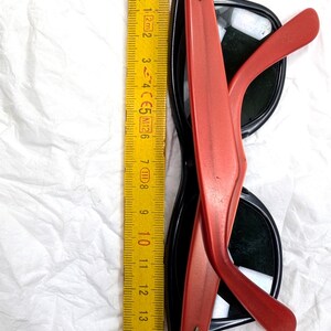 May include: Black sunglasses with red arms, measuring approximately 6 inches in length. The sunglasses have a classic design with dark lenses and a contrasting color scheme. The image also includes a yellow ruler.