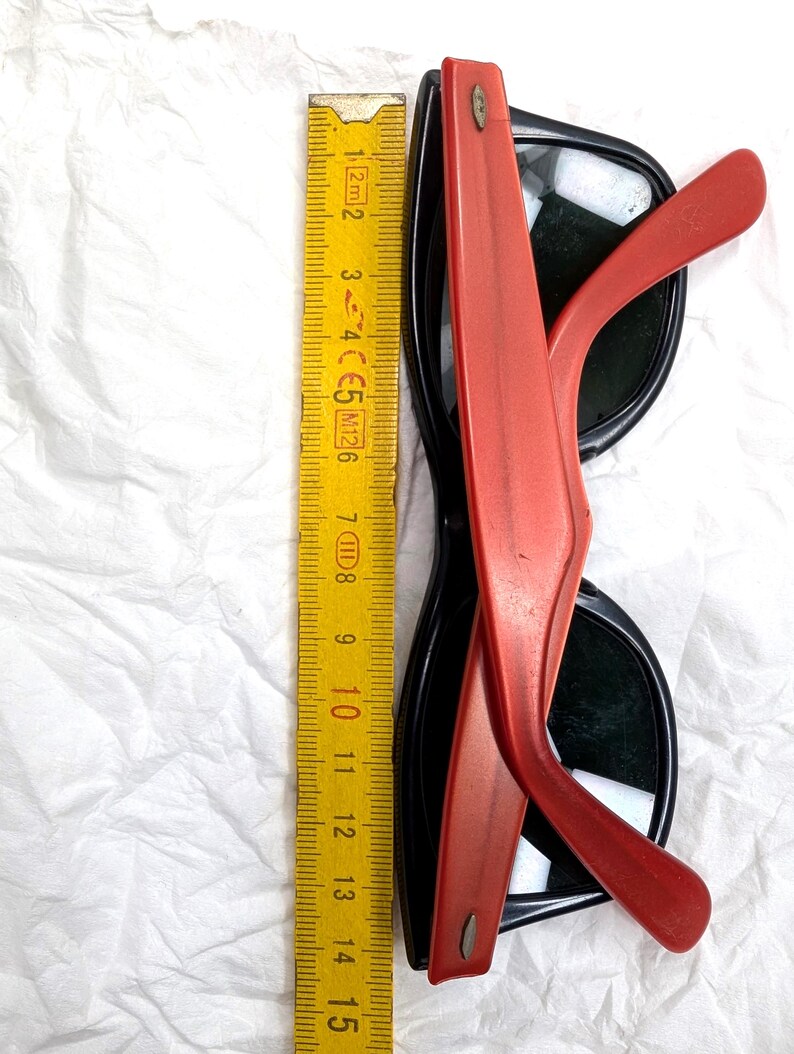 May include: Black sunglasses with red arms, measuring approximately 6 inches in length. The sunglasses have a classic design with dark lenses and a contrasting color scheme. The image also includes a yellow ruler.