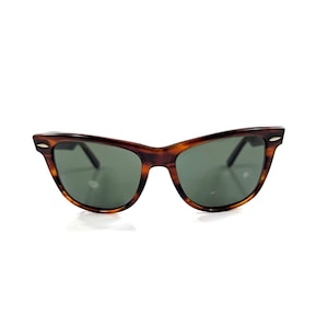 Op de afbeelding: Schildpadkleurige cat-eye zonnebril met een bruin en zwart gevlekt montuur en donkergroen getinte glazen. Het montuur is voorzien van kleine zilverkleurige metalen accenten op de buitenste hoeken van het voorstuk voor een klassieke uitstraling.