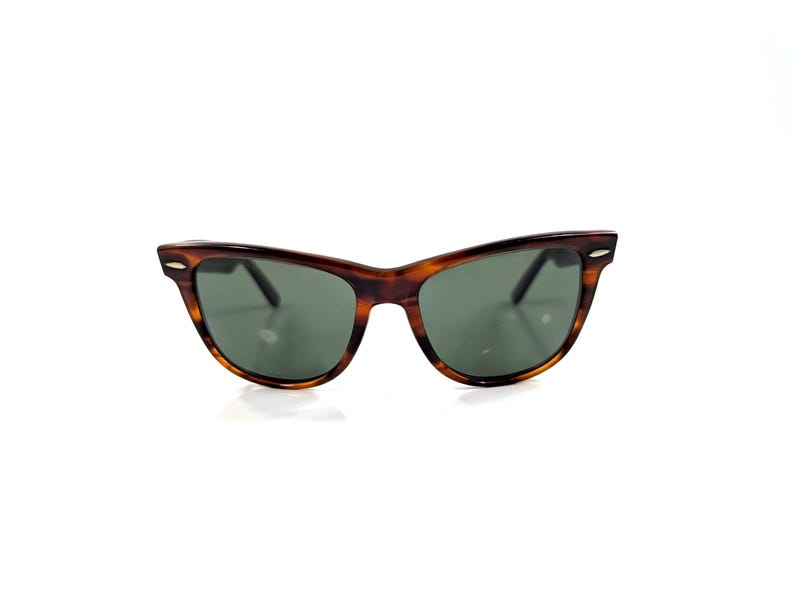 Op de afbeelding: Schildpadkleurige cat-eye zonnebril met een bruin en zwart gevlekt montuur en donkergroen getinte glazen. Het montuur is voorzien van kleine zilverkleurige metalen accenten op de buitenste hoeken van het voorstuk voor een klassieke uitstraling.