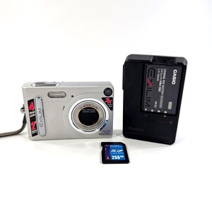 Op de afbeelding: Een zilveren Casio Exilim digitale camera met een 3x optische zoomlens, een zwarte acculader met het opschrift Casio Exilim en een blauwe 256MB SD-geheugenkaart. De camera is voorzien van een 4,0 megapixel sensor en een grijze polsband.