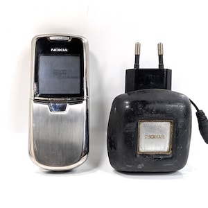 Op de afbeelding: Een zilveren Nokia mobiele telefoon en een zwarte Nokia oplader. Het scherm van de telefoon toont de tijd. De oplader heeft een zilveren vierkant met het Nokia-logo. De telefoon heeft een metallic afwerking.
