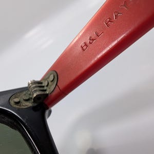 May include: Close-up of a pair of sunglasses with black frames and red temples. The text "B&L RAY-BAN" is embossed on the red temple. The lenses are green.