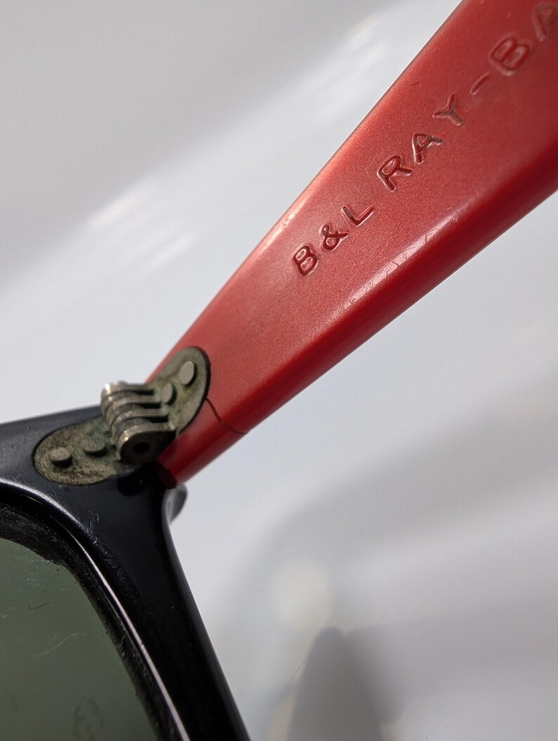 May include: Close-up of a pair of sunglasses with black frames and red temples. The text "B&L RAY-BAN" is embossed on the red temple. The lenses are green.