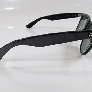 Op de afbeelding: Zwarte zonnebril met een textuurframe met visgraatpatroon en donkere glazen. De pootjes zijn voorzien van het goudkleurige Ray-Ban logo bij de scharnieren. Het montuur heeft een matte afwerking en toont duidelijk het zijprofiel.