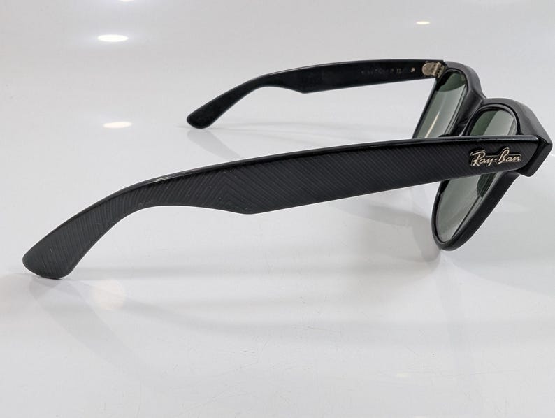 Op de afbeelding: Zwarte zonnebril met een textuurframe met visgraatpatroon en donkere glazen. De pootjes zijn voorzien van het goudkleurige Ray-Ban logo bij de scharnieren. Het montuur heeft een matte afwerking en toont duidelijk het zijprofiel.