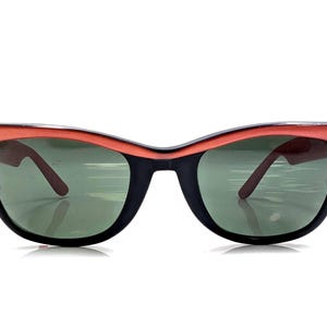 May include: Vintage cat-eye sunglasses with a black frame and coral-colored top rim. The lenses are green, and the arms are a dark brown. These retro sunglasses offer a stylish look.