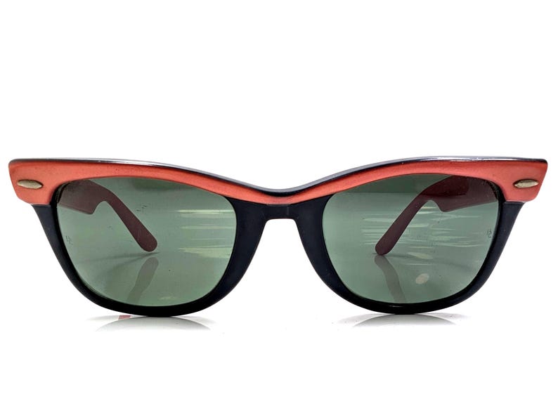 May include: Vintage cat-eye sunglasses with a black frame and coral-colored top rim. The lenses are green, and the arms are a dark brown. These retro sunglasses offer a stylish look.