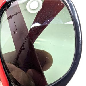 May include: Close-up of a pair of sunglasses with a black frame and red arms. The lenses are green and have a dark, reflective coating. The sunglasses appear to be vintage or retro style.