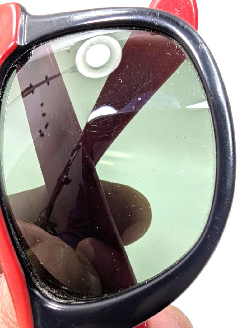 May include: Close-up of a pair of sunglasses with a black frame and red arms. The lenses are green and have a dark, reflective coating. The sunglasses appear to be vintage or retro style.