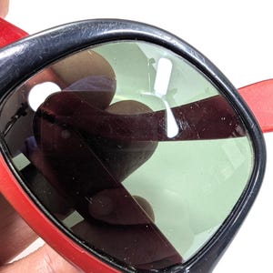 May include: Close-up of sunglasses with a red frame and temples, and black rims. The lenses are a light green color. The sunglasses are held by a person's hand.