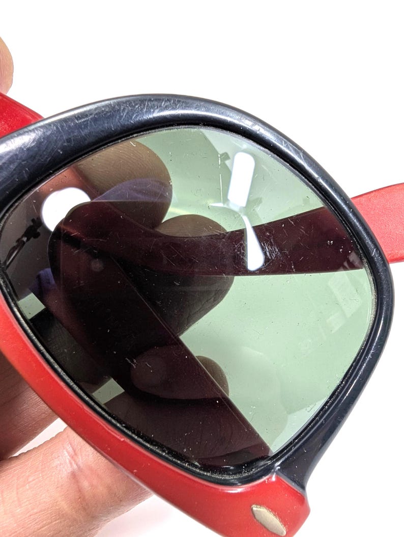 May include: Close-up of sunglasses with a red frame and temples, and black rims. The lenses are a light green color. The sunglasses are held by a person's hand.