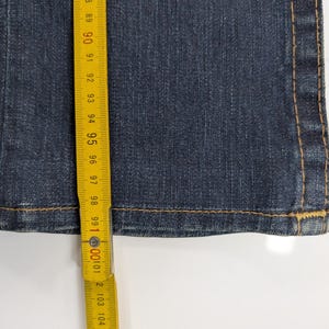 Pode incluir: Close-up de jeans de ganga azul escuro com costuras amarelas vis&iacute;veis. Uma fita m&eacute;trica amarela mostra o comprimento dos jeans. Os jeans t&ecirc;m cerca de 100 cm de comprimento.