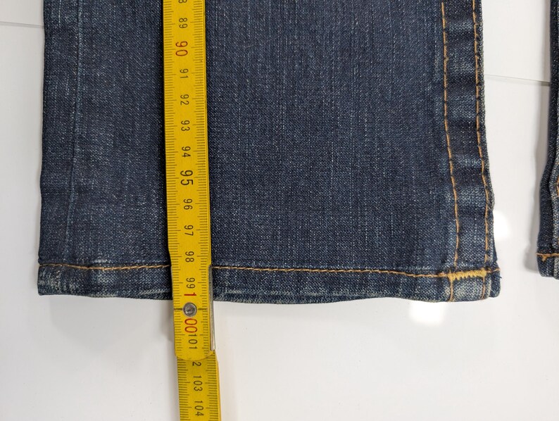 Pode incluir: Close-up de jeans de ganga azul escuro com costuras amarelas vis&iacute;veis. Uma fita m&eacute;trica amarela mostra o comprimento dos jeans. Os jeans t&ecirc;m cerca de 100 cm de comprimento.