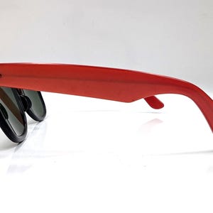 May include: Red sunglasses with black frames and dark lenses. The sunglasses have a classic, retro design with a thick, rectangular frame and a curved temple arm. The sunglasses are shown from a side view.