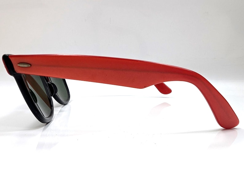 May include: Red sunglasses with black frames and dark lenses. The sunglasses have a classic, retro design with a thick, rectangular frame and a curved temple arm. The sunglasses are shown from a side view.