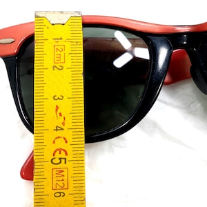 May include: Black sunglasses with red frames. The sunglasses are measured with a yellow ruler. The sunglasses have dark lenses and a classic design. The frame measures approximately 2 inches.
