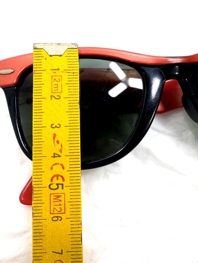 May include: Black sunglasses with red frames. The sunglasses are measured with a yellow ruler. The sunglasses have dark lenses and a classic design. The frame measures approximately 2 inches.