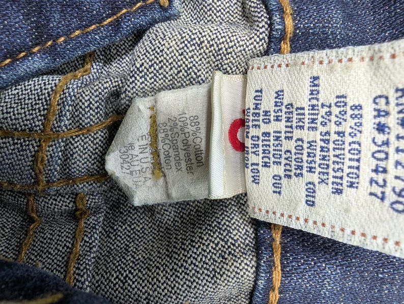 Pode incluir: Vista de perto do interior de uma pe&ccedil;a de roupa de ganga, mostrando costuras e etiquetas de tecido. As etiquetas exibem detalhes da composi&ccedil;&atilde;o do tecido: 88% algod&atilde;o, 10% poli&eacute;ster e 2% spandex. Instru&ccedil;&otilde;es de lavagem vis&iacute;veis.