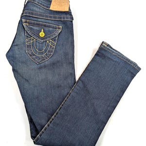 Pode incluir: Jeans de ganga azul escuro com um design cl&aacute;ssico de cinco bolsos. Apresenta um bolso traseiro decorativo com costura amarela e um remendo de couro. Os jeans est&atilde;o dispostos sobre uma superf&iacute;cie branca.