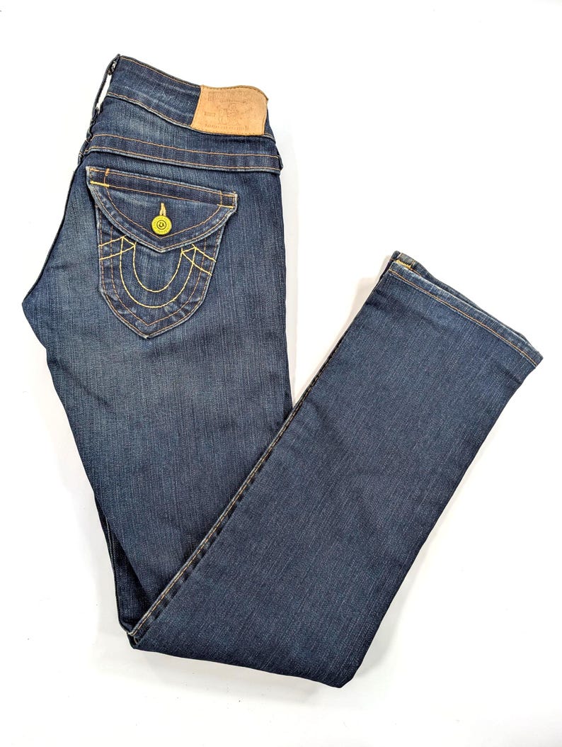 Pode incluir: Jeans de ganga azul escuro com um design cl&aacute;ssico de cinco bolsos. Apresenta um bolso traseiro decorativo com costura amarela e um remendo de couro. Os jeans est&atilde;o dispostos sobre uma superf&iacute;cie branca.