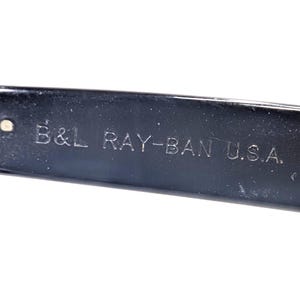 Op de afbeelding: Close-up van een zwarte brilpoot met de gegraveerde tekst "B&L RAY-BAN U.S.A.". De tekst is in een schreefloze lettertype. De poot heeft een glanzend, reflecterend oppervlak.