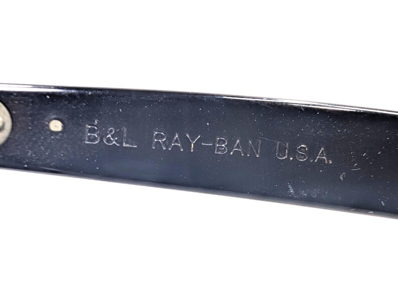 Op de afbeelding: Close-up van een zwarte brilpoot met de gegraveerde tekst "B&L RAY-BAN U.S.A.". De tekst is in een schreefloze lettertype. De poot heeft een glanzend, reflecterend oppervlak.