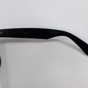 Op de afbeelding: Zwarte zonnebril met een gebogen brilpoot, voorzien van een metalen scharnier en de in reli&euml;f aangebrachte tekst B&L RAY-BAN U.S.A. aan de binnenzijde. Het montuur is matzwart met een donkergroene lens zichtbaar aan de linkerkant.