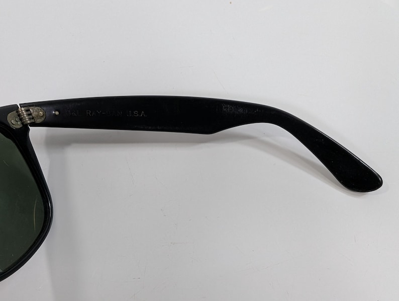 Op de afbeelding: Zwarte zonnebril met een gebogen brilpoot, voorzien van een metalen scharnier en de in reli&euml;f aangebrachte tekst B&L RAY-BAN U.S.A. aan de binnenzijde. Het montuur is matzwart met een donkergroene lens zichtbaar aan de linkerkant.