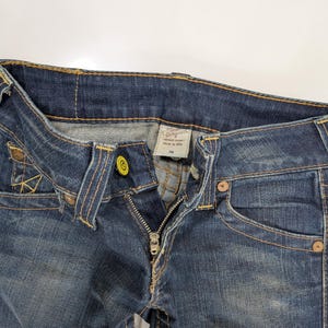 Pode incluir: Close-up de jeans de ganga azul com fecho parcialmente aberto. Os jeans t&ecirc;m um design cl&aacute;ssico de cinco bolsos, com costuras vis&iacute;veis em fio amarelo contrastante. A cintura apresenta um fecho de bot&atilde;o e uma etiqueta.