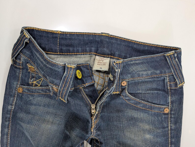 Pode incluir: Close-up de jeans de ganga azul com fecho parcialmente aberto. Os jeans t&ecirc;m um design cl&aacute;ssico de cinco bolsos, com costuras vis&iacute;veis em fio amarelo contrastante. A cintura apresenta um fecho de bot&atilde;o e uma etiqueta.