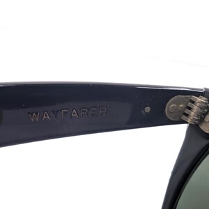 Op de afbeelding: Close-up van een zwarte Wayfarer zonnebril. De tekst "WAYFARER" is in reli&euml;f op de poot. Het scharnier is zichtbaar, met een metallic afwerking. De lenzen zijn donkergroen.