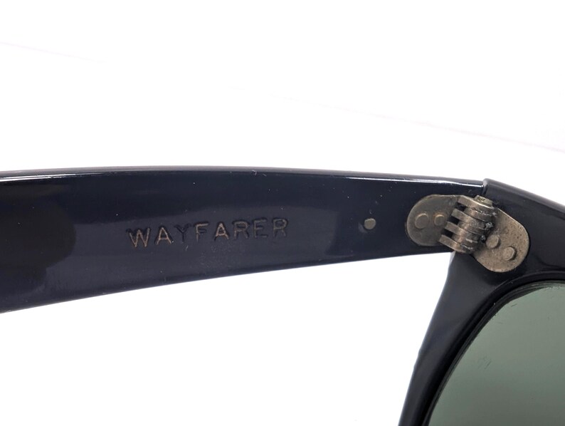 Op de afbeelding: Close-up van een zwarte Wayfarer zonnebril. De tekst "WAYFARER" is in reli&euml;f op de poot. Het scharnier is zichtbaar, met een metallic afwerking. De lenzen zijn donkergroen.