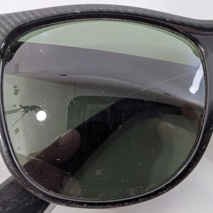 Op de afbeelding: Zwarte zonnebril met een dik kunststof montuur, een textuurpatroon op de pootjes en een klein metalen accent bij het scharnier. De donkergroene glazen vertonen zichtbare krassen op het oppervlak en reflecties in het glas.