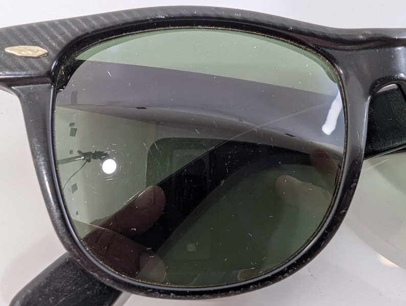 Op de afbeelding: Zwarte zonnebril met een dik kunststof montuur, een textuurpatroon op de pootjes en een klein metalen accent bij het scharnier. De donkergroene glazen vertonen zichtbare krassen op het oppervlak en reflecties in het glas.