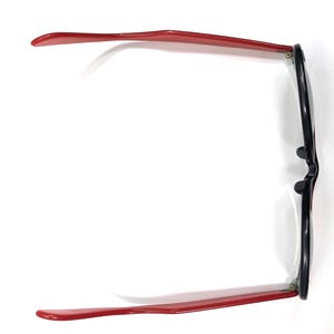 May include: Overhead view of eyeglasses with a black frame and red temples. The glasses are set against a white background, showcasing their design. The red temples extend outwards, providing a striking contrast to the black frame.