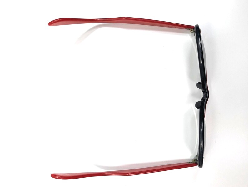 May include: Overhead view of eyeglasses with a black frame and red temples. The glasses are set against a white background, showcasing their design. The red temples extend outwards, providing a striking contrast to the black frame.