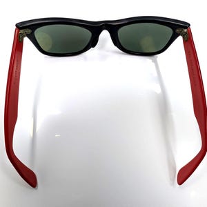 May include: Black sunglasses with red arms. The sunglasses have a classic design with a black frame and green lenses. The red arms have text printed on them.