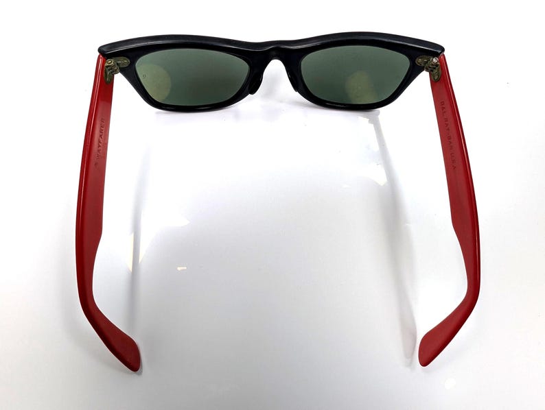 May include: Black sunglasses with red arms. The sunglasses have a classic design with a black frame and green lenses. The red arms have text printed on them.