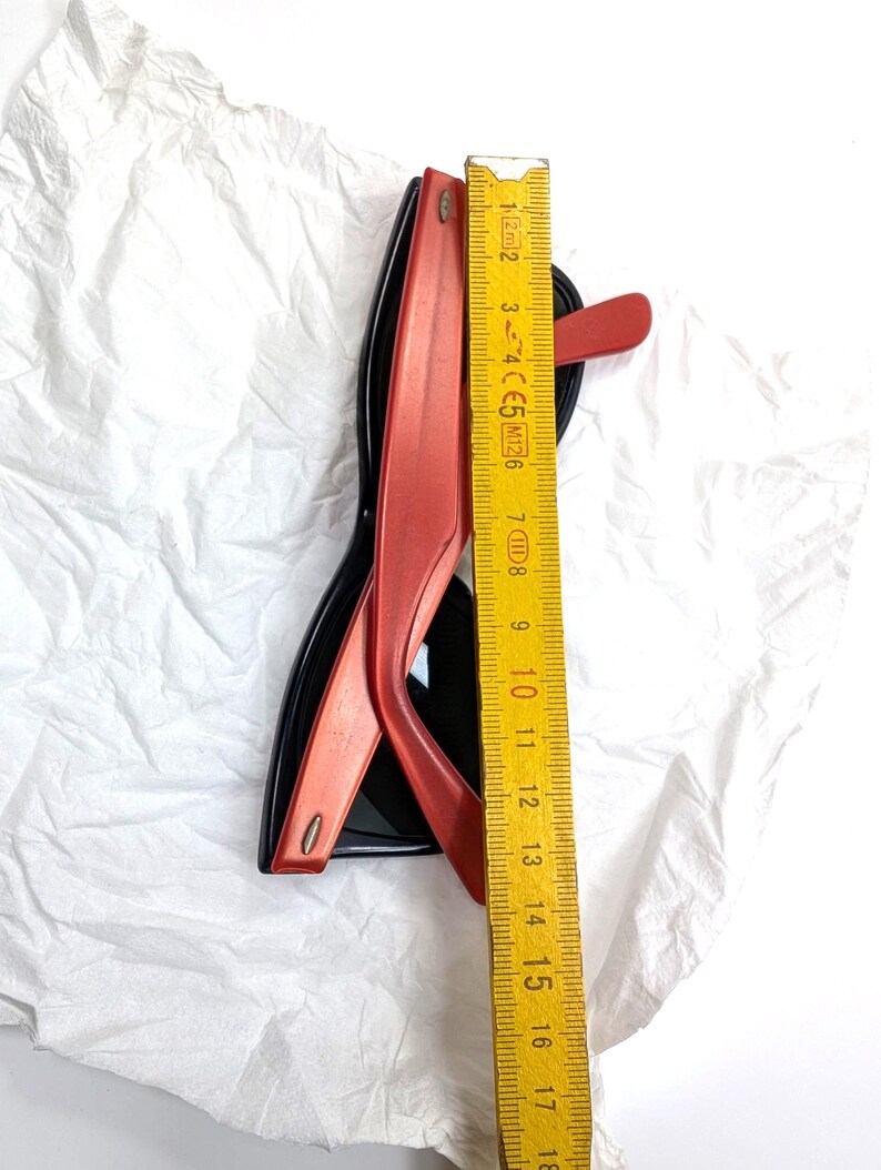 May include: Black and red sunglasses with a ruler showing a measurement of approximately 6 inches. The sunglasses have a classic design with a black frame and red arms. The ruler is yellow with black markings.
