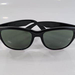 Op de afbeelding: Zwarte zonnebril in wayfarer-stijl met donkergroen getinte glazen en een glanzend zwart montuur. Het montuur is voorzien van zilverkleurige metalen accenten op de buitenste hoeken, verbonden met de pootjes via een klassiek scharnierontwerp.