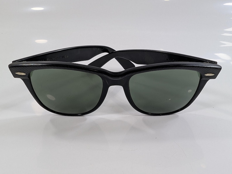 Op de afbeelding: Zwarte zonnebril in wayfarer-stijl met donkergroen getinte glazen en een glanzend zwart montuur. Het montuur is voorzien van zilverkleurige metalen accenten op de buitenste hoeken, verbonden met de pootjes via een klassiek scharnierontwerp.