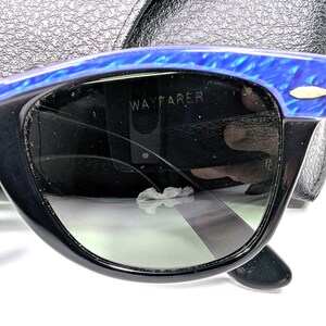 Op de afbeelding: Zwarte Wayfarer zonnebril met blauwe gemarmerde accenten op het bovenste montuur. De donkere glazen tonen het woord "WAYFARER". De zonnebril ligt op een zwart leren etui.