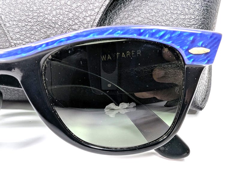 Op de afbeelding: Zwarte Wayfarer zonnebril met blauwe gemarmerde accenten op het bovenste montuur. De donkere glazen tonen het woord "WAYFARER". De zonnebril ligt op een zwart leren etui.