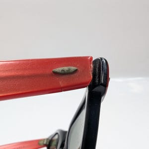May include: Close-up of vintage eyeglasses with a red frame and black arm. The red frame shows signs of wear, with a small crack visible. The black arm is attached to the red frame with a metal hinge.