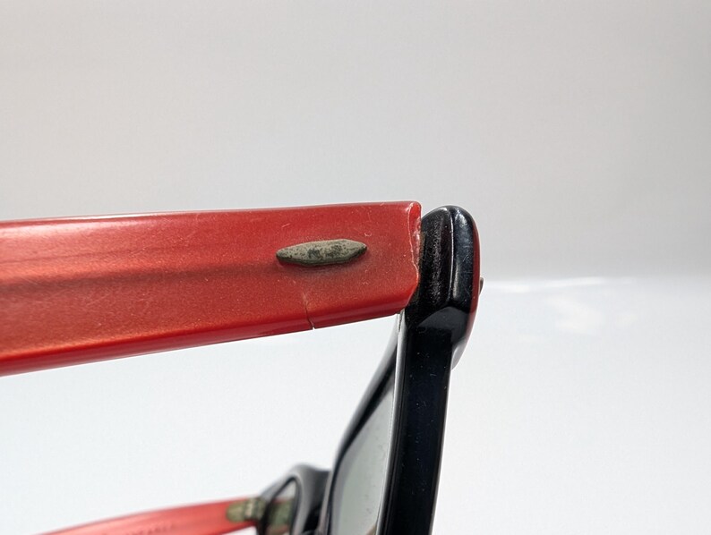 May include: Close-up of vintage eyeglasses with a red frame and black arm. The red frame shows signs of wear, with a small crack visible. The black arm is attached to the red frame with a metal hinge.