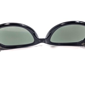Op de afbeelding: Zwarte zonnebril met een cat-eye vorm. De monturen zijn glanzend en de glazen zijn groen getint. De scharnieren zijn zilverkleurig. Op een witte achtergrond.