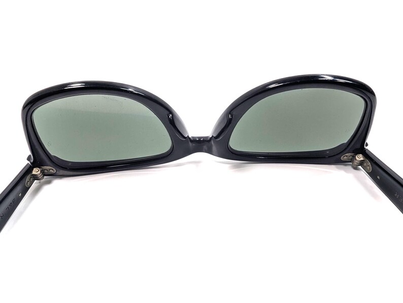 Op de afbeelding: Zwarte zonnebril met een cat-eye vorm. De monturen zijn glanzend en de glazen zijn groen getint. De scharnieren zijn zilverkleurig. Op een witte achtergrond.
