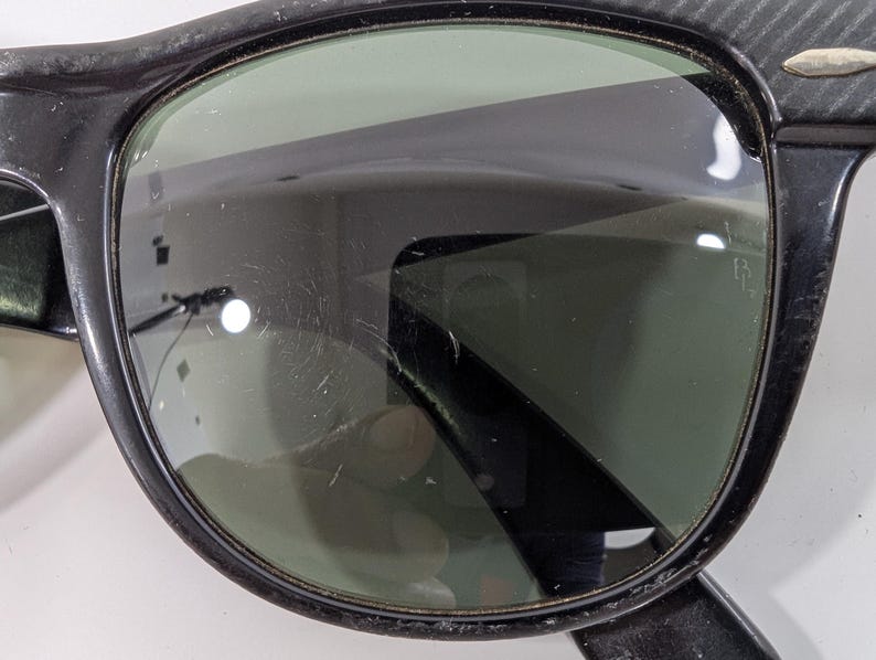 Op de afbeelding: Zwarte zonnebril met een dik kunststof montuur met een textuurpatroon op de bovenhoeken en veren. De glazen zijn donkergroen en vertonen zichtbare krassen op het oppervlak. Een klein zilverkleurig metalen accent is zichtbaar op de hoek van het montuur.